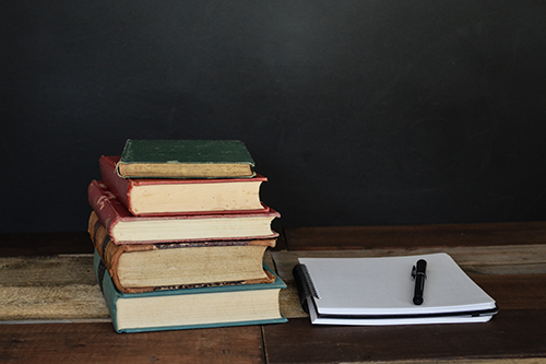 A wooden desk with a stack of hard cover books and a spiral-bound pad of paper with a pen on top