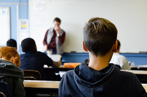 An image showing the back of students' heads in a classroom with a teacher at the whiteboard