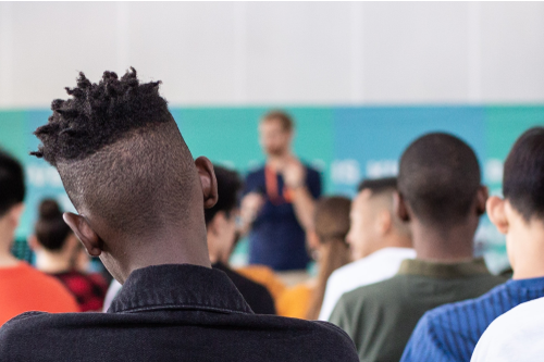 This photo shows the backs of high school students' heads as they are in a class and watching their teacher in the front of the room