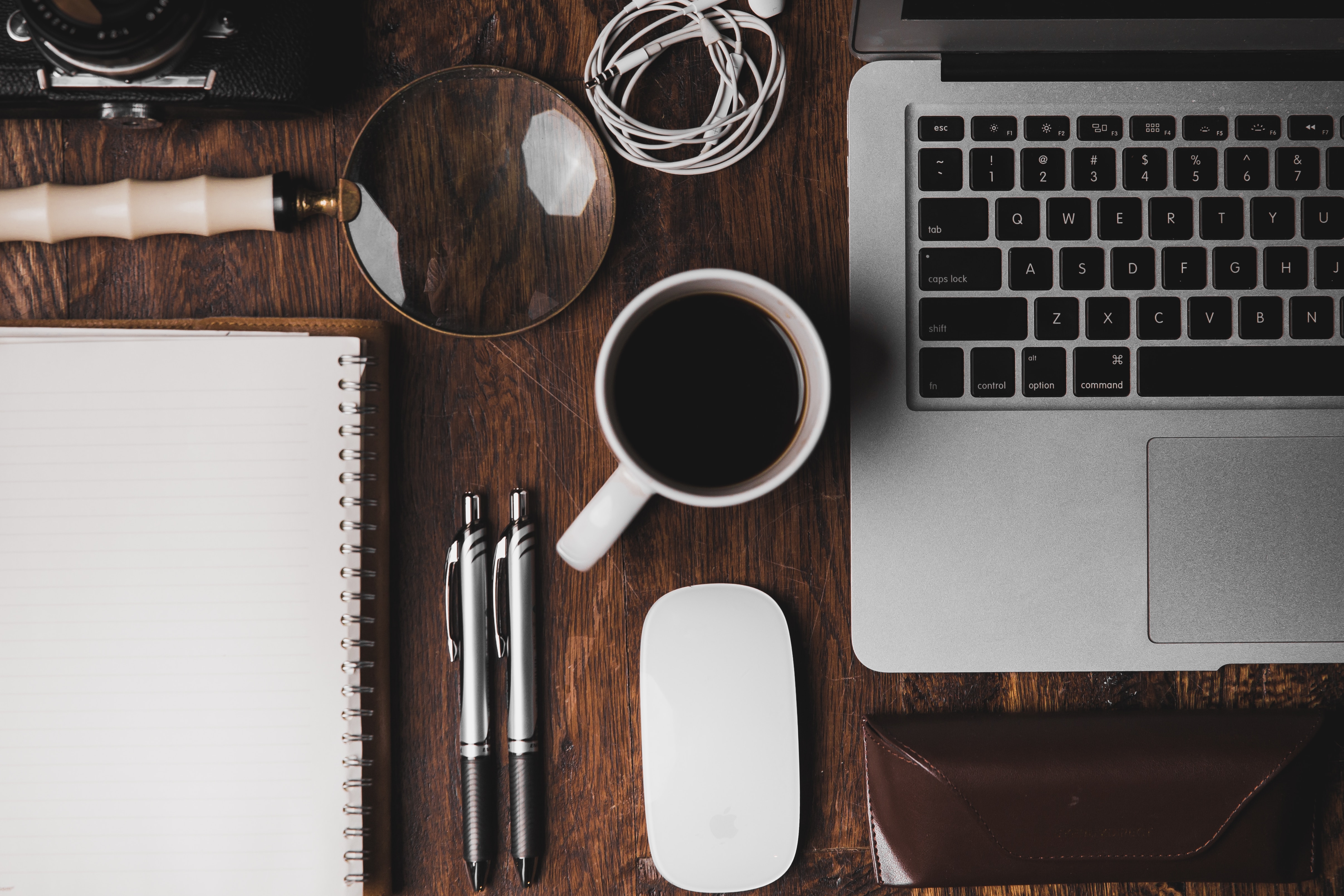 A desk top crowded with notepad, pens, coffee mug, glasses case, and a laptop computer